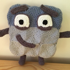 Könnte beinhalten: Ein gehäkeltes Plüschtier in Form eines Quadrats in Blau- und Grautönen. Das Spielzeug hat ein lächelndes Gesicht mit braunen Augenbrauen, weißen Augen und einem braunen Mund.