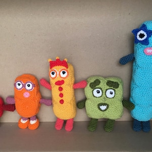 Könnte beinhalten: Fünf gehäkelte Stofftiere in einem Karton. Die Spielzeuge sind in verschiedenen Farben und Formen, darunter ein roter Würfel, ein orangefarbenes Rechteck, ein gelbes Rechteck mit einer Krone, ein grünes Quadrat und ein blaues Rechteck mit einer Sonnenbrille.