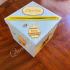 May include: A light blue card box with a yellow trim and a Winnie the Pooh theme. The box has a slot for cards and a label that says "Cards".