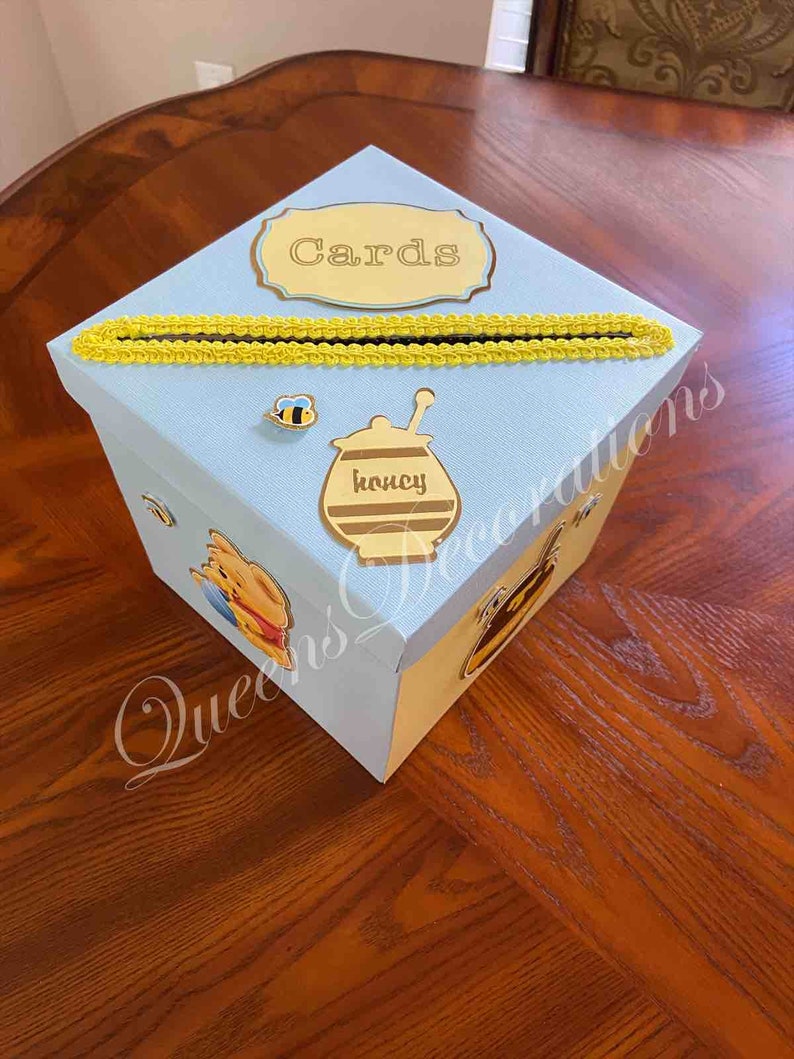 May include: A light blue card box with a yellow trim and a Winnie the Pooh theme. The box has a slot for cards and a label that says "Cards".