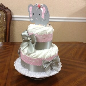 May include: A two-tier diaper cake decorated with pink and white polka dot ribbon, silver ribbon, and a silver elephant topper with a pink flower.