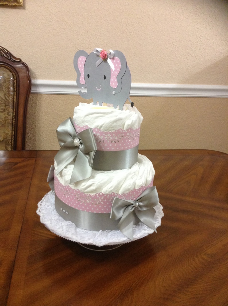 May include: A two-tier diaper cake decorated with pink and white polka dot ribbon, silver ribbon, and a silver elephant topper with a pink flower.