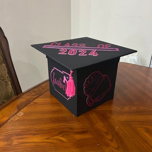 K&ouml;nnte beinhalten: Schwarze Abschluss-Hut-Box mit rosa Verzierungen und dem Text "Class of 2024". Die Box hat einen rosa Quasten und eine rosa Megaphon-Grafik.