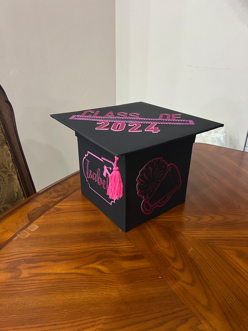 K&ouml;nnte beinhalten: Schwarze Abschluss-Hut-Box mit rosa Verzierungen und dem Text "Class of 2024". Die Box hat einen rosa Quasten und eine rosa Megaphon-Grafik.