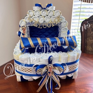 Puede incluir: Un pastel de pa&ntilde;ales azul y dorado con forma de carruaje. El pastel est&aacute; decorado con una cinta azul, purpurina dorada y una peque&ntilde;a figura de pr&iacute;ncipe. El pastel est&aacute; sobre una mesa de madera.