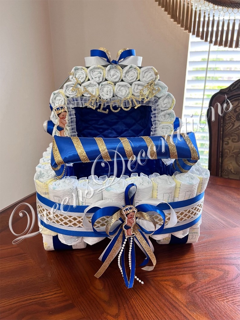 Puede incluir: Un pastel de pa&ntilde;ales azul y dorado con forma de carruaje. El pastel est&aacute; decorado con una cinta azul, purpurina dorada y una peque&ntilde;a figura de pr&iacute;ncipe. El pastel est&aacute; sobre una mesa de madera.