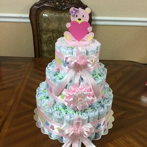 May include: A three-tiered nappy cake decorated with pink and white ribbons, a pink bow, and a pink teddy bear holding a pink heart. The cake is on a wooden table.