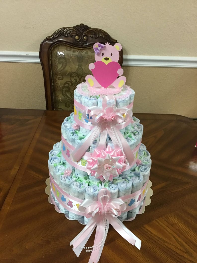 May include: A three-tiered nappy cake decorated with pink and white ribbons, a pink bow, and a pink teddy bear holding a pink heart. The cake is on a wooden table.