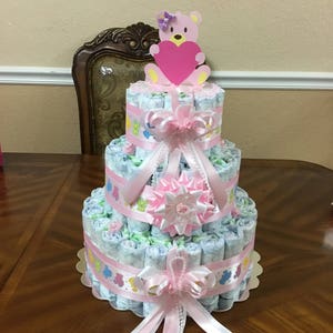 May include: A three-tiered nappy cake decorated with pink and white ribbons, a pink bow, and a pink teddy bear holding a pink heart. The cake is on a wooden table.