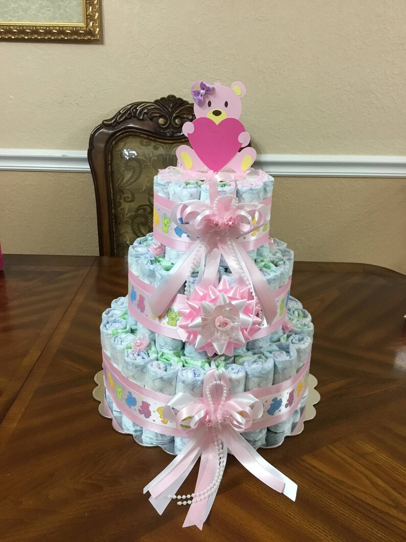 May include: A three-tiered nappy cake decorated with pink and white ribbons, a pink bow, and a pink teddy bear holding a pink heart. The cake is on a wooden table.