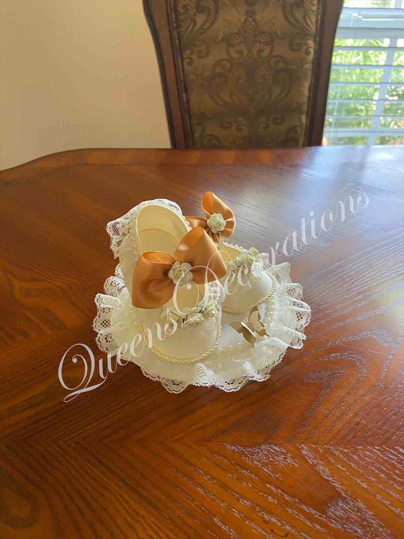May include: Two white baby shoes with lace trim and orange satin bows. The shoes are on a white lace pillow.