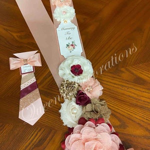 May include: A pink satin sash with a white tag that says "Mommy To Be" and a burgundy, gold, and white floral arrangement. A matching pink bow tie with a burgundy and gold striped pattern says "Daddy To Be".