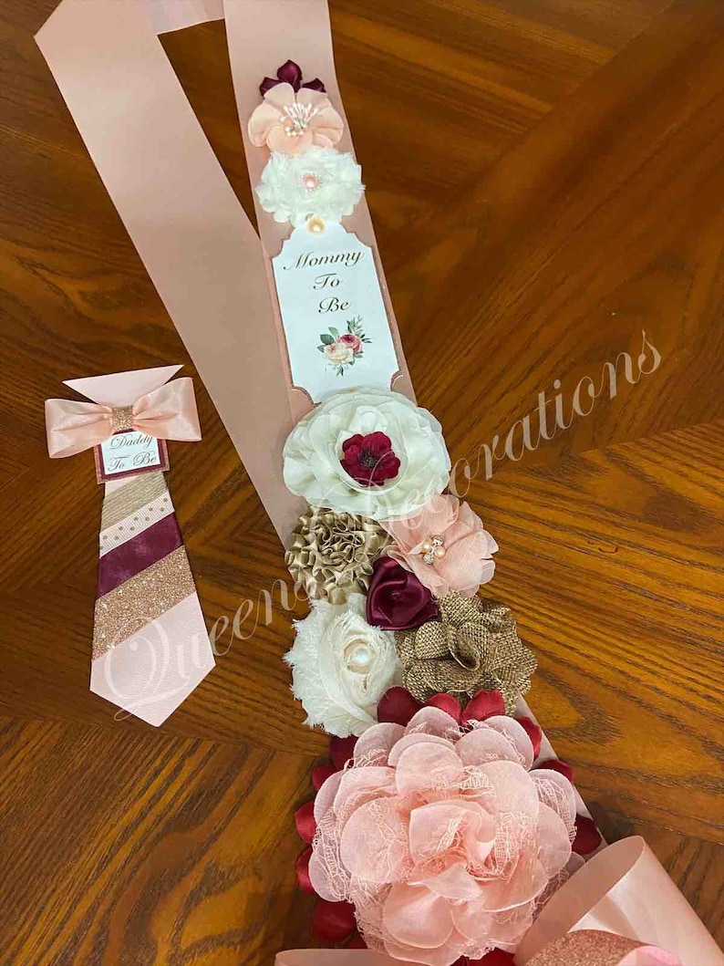 May include: A pink satin sash with a white tag that says "Mommy To Be" and a burgundy, gold, and white floral arrangement. A matching pink bow tie with a burgundy and gold striped pattern says "Daddy To Be".