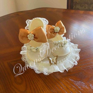 May include: A pair of white baby shoes with gold bows and white flowers on a white lace base with a gold butterfly.