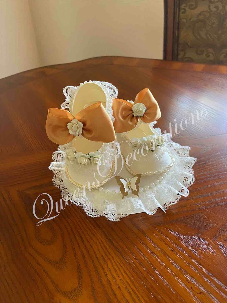 May include: A pair of white baby shoes with gold bows and white flowers on a white lace base with a gold butterfly.