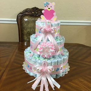 May include: A three-tiered nappy cake decorated with pink and white ribbons, a pink teddy bear topper, and a pink and white bow. The cake is made of rolled nappies and decorated with various baby-themed embellishments.