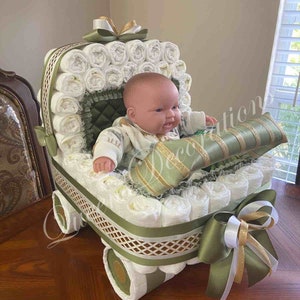 Puede incluir: Un pastel de pa&ntilde;ales con forma de cochecito de beb&eacute; con una mu&ntilde;eca sentada dentro. El cochecito est&aacute; decorado con cinta verde y dorada y un lazo blanco. La mu&ntilde;eca lleva un atuendo a rayas verde y blanco.