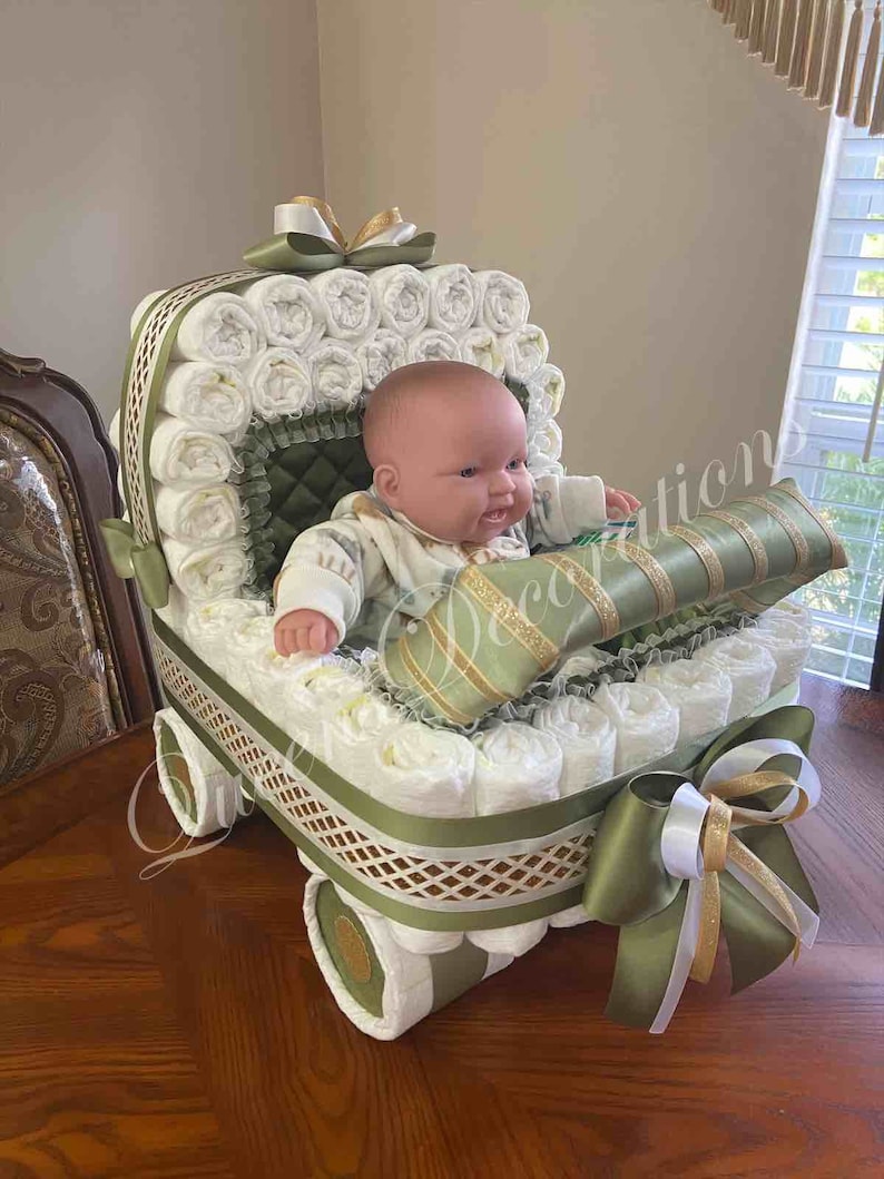 Puede incluir: Un pastel de pa&ntilde;ales con forma de cochecito de beb&eacute; con una mu&ntilde;eca sentada dentro. El cochecito est&aacute; decorado con cinta verde y dorada y un lazo blanco. La mu&ntilde;eca lleva un atuendo a rayas verde y blanco.