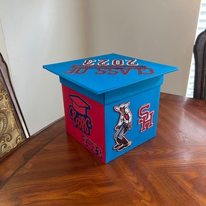 K&ouml;nnte beinhalten: Eine blaue und rote Abschlussbox mit einem Abschlusskappen-Deckel. Die Box hat den Text "Class of 2023" auf dem Deckel und einen roten und wei&szlig;en "Class of 2023" Aufkleber an der Seite. Es gibt einen Cowboy-Aufkleber und einen roten und wei&szlig;en "SH" Aufkleber auf der Box.