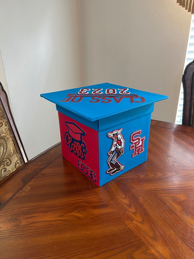 K&ouml;nnte beinhalten: Eine blaue und rote Abschlussbox mit einem Abschlusskappen-Deckel. Die Box hat den Text "Class of 2023" auf dem Deckel und einen roten und wei&szlig;en "Class of 2023" Aufkleber an der Seite. Es gibt einen Cowboy-Aufkleber und einen roten und wei&szlig;en "SH" Aufkleber auf der Box.