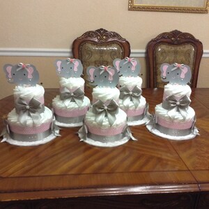 May include: Five gray and pink diaper cakes decorated with elephant toppers. Each cake has two tiers and is wrapped in white lace. The cakes are arranged on a wooden table.