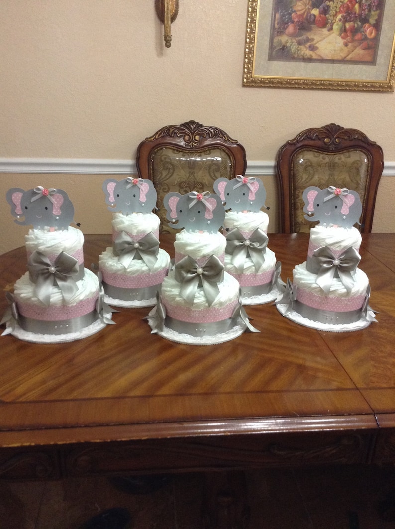 May include: Five gray and pink diaper cakes decorated with elephant toppers. Each cake has two tiers and is wrapped in white lace. The cakes are arranged on a wooden table.