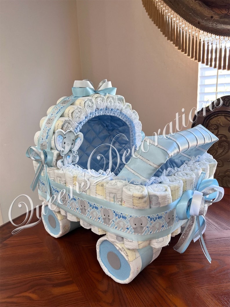 May include: A blue and white diaper cake shaped like a baby carriage. The carriage is decorated with a blue ribbon, a white lace trim, and a small stuffed elephant. The wheels of the carriage are made of rolled up diapers.