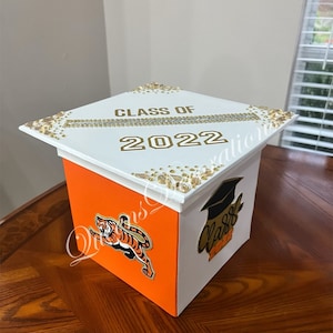 May include: White graduation cap box with orange sides. The top of the box has gold glitter and the text "CLASS OF 2022". One side of the box has a tiger mascot and the other side has a graduation cap with the text "Class of 2022".