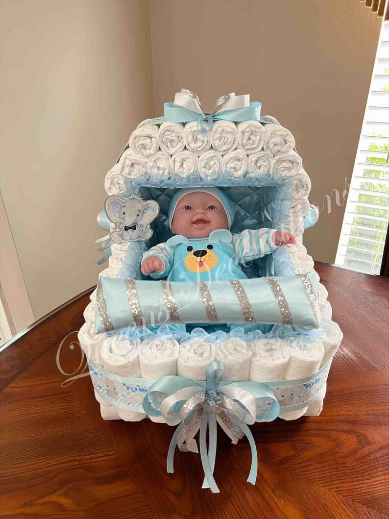 May include: A blue and white diaper cake shaped like a baby carriage with a baby doll inside. The carriage is decorated with blue and white ribbons and a silver glitter pillow.