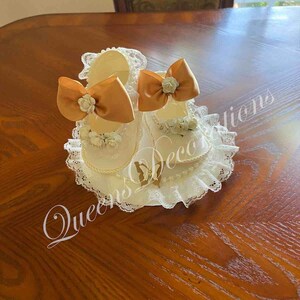 May include: Two white baby shoes with gold bows and lace trim. The shoes are on a white lace doily with a gold butterfly design. The text "Queen's Decorations" is visible in the image.