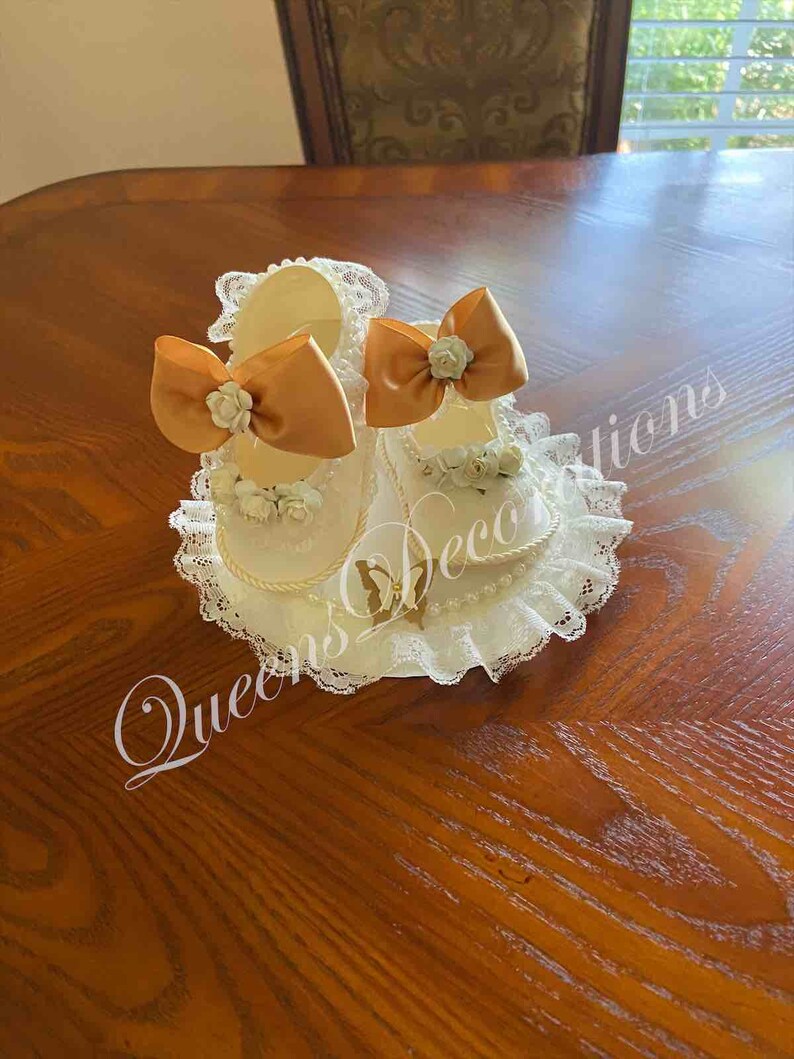 May include: Two white baby shoes with gold bows and lace trim. The shoes are on a white lace doily with a gold butterfly design. The text "Queen's Decorations" is visible in the image.
