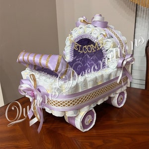 Puede incluir: Un pastel de pa&ntilde;ales morado y dorado con forma de cochecito. El cochecito est&aacute; decorado con cinta blanca y dorada, y las palabras "Bienvenido" est&aacute;n escritas en letras doradas.
