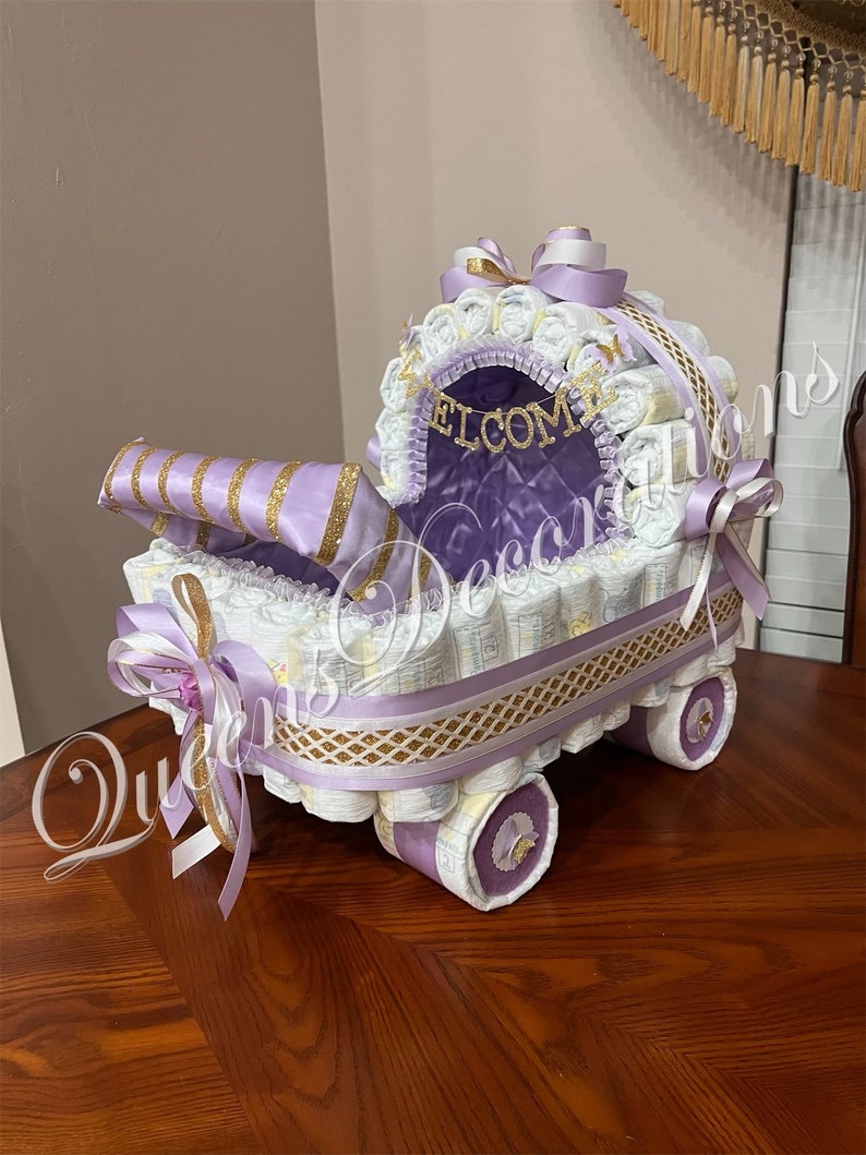 Puede incluir: Un pastel de pa&ntilde;ales morado y dorado con forma de cochecito. El cochecito est&aacute; decorado con cinta blanca y dorada, y las palabras "Bienvenido" est&aacute;n escritas en letras doradas.