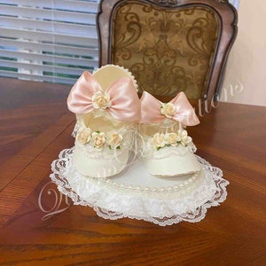 May include: Two white baby shoes with pink bows and lace trim, displayed on a white lace doily with pearl accents.