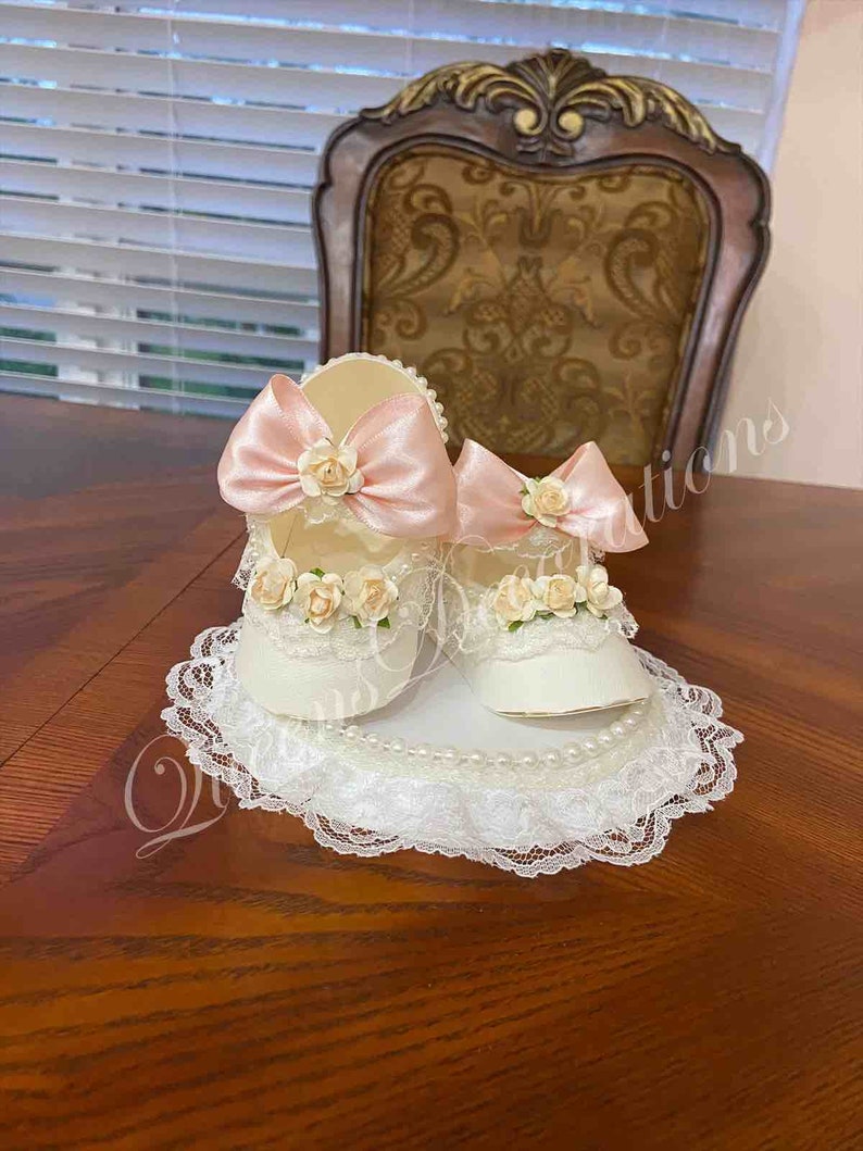 May include: Two white baby shoes with pink bows and lace trim, displayed on a white lace doily with pearl accents.