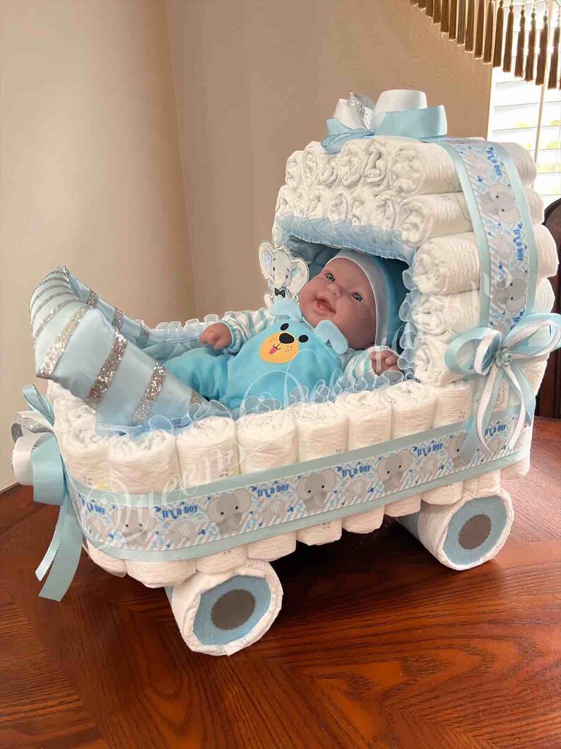 May include: A blue and white diaper cake shaped like a baby carriage with a baby doll inside. The carriage is decorated with a blue ribbon and a gray ribbon with the words "It's a Boy" printed on it. The wheels are made of rolled up diapers.