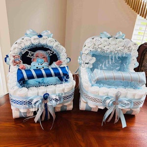 Puede incluir: Dos carritos de pastel de pa&ntilde;ales azules y blancos decorados con cinta azul y plateada. Un carrito tiene un mu&ntilde;eco.