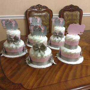 May include: Four gray and pink diaper cakes decorated with elephant toppers. Each cake has a silver ribbon and a white lace base.