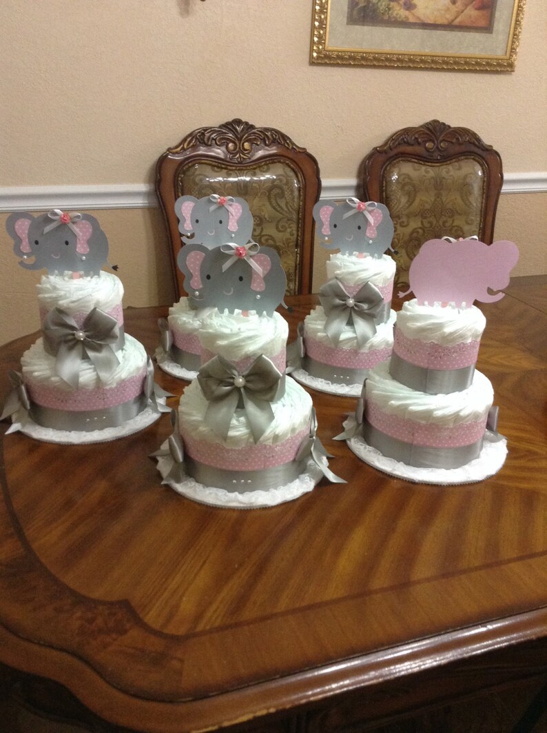 May include: Four gray and pink diaper cakes decorated with elephant toppers. Each cake has a silver ribbon and a white lace base.