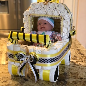 Puede incluir: Un pastel de pa&ntilde;ales en forma de cochecito de beb&eacute; amarillo y negro con una mu&ntilde;eca de beb&eacute; dentro. El cochecito est&aacute; decorado con cinta amarilla y negra y un amuleto de abeja.