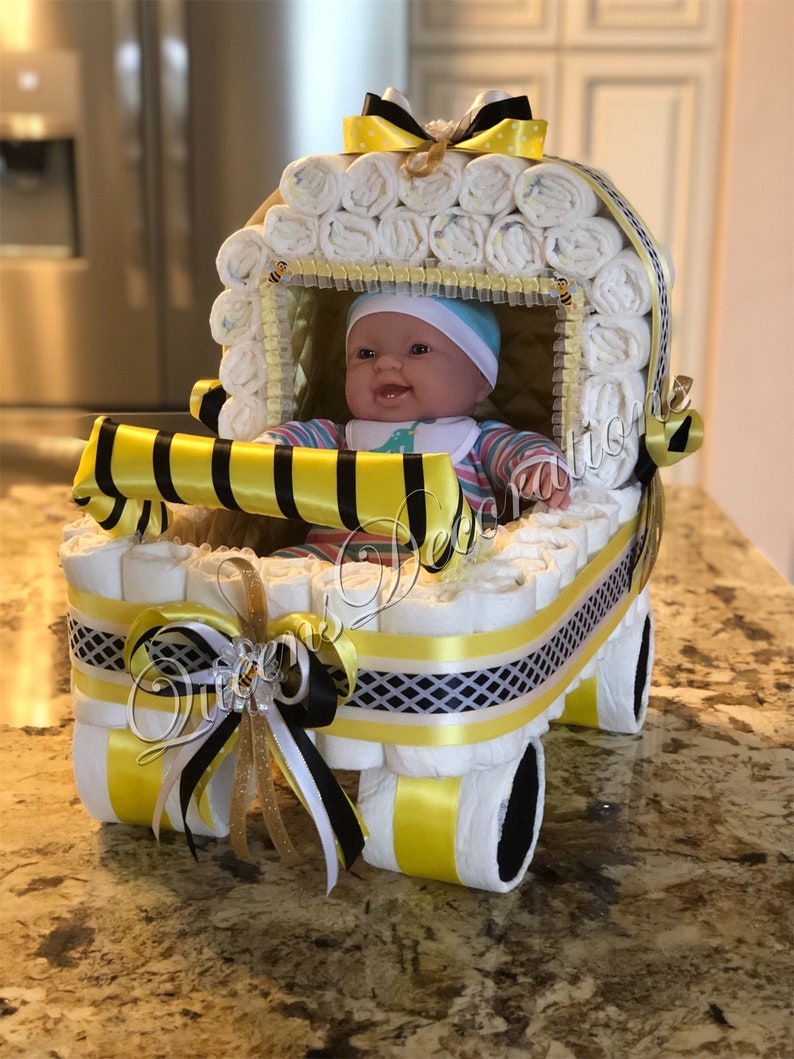 Puede incluir: Un pastel de pa&ntilde;ales en forma de cochecito de beb&eacute; amarillo y negro con una mu&ntilde;eca de beb&eacute; dentro. El cochecito est&aacute; decorado con cinta amarilla y negra y un amuleto de abeja.