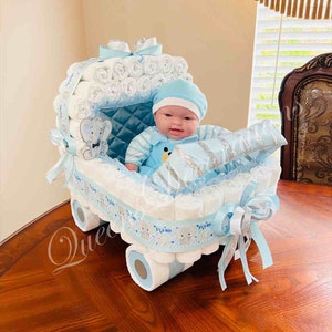 May include: A blue and white diaper cake shaped like a baby carriage with a baby doll sitting inside. The carriage is decorated with a blue ribbon and a silver ribbon. The baby doll is wearing a blue and white striped outfit and a blue hat.