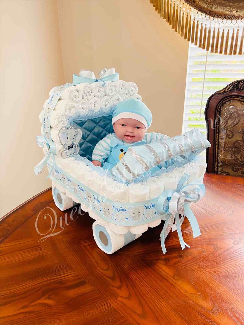 May include: A blue and white diaper cake shaped like a baby carriage with a baby doll sitting inside. The carriage is decorated with a blue ribbon and a silver ribbon. The baby doll is wearing a blue and white striped outfit and a blue hat.