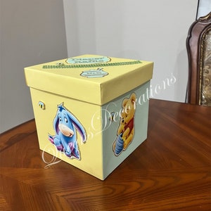 May include: A yellow and blue diaper raffle box with Winnie the Pooh and Eeyore characters. The box has a green ribbon and the text "Diaper Raffle" on the top.