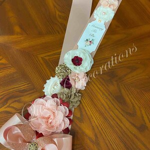 May include: A pink satin sash with a white card that says "Mommy To Be" and a floral design. The sash is decorated with burgundy, blush, and gold flowers.