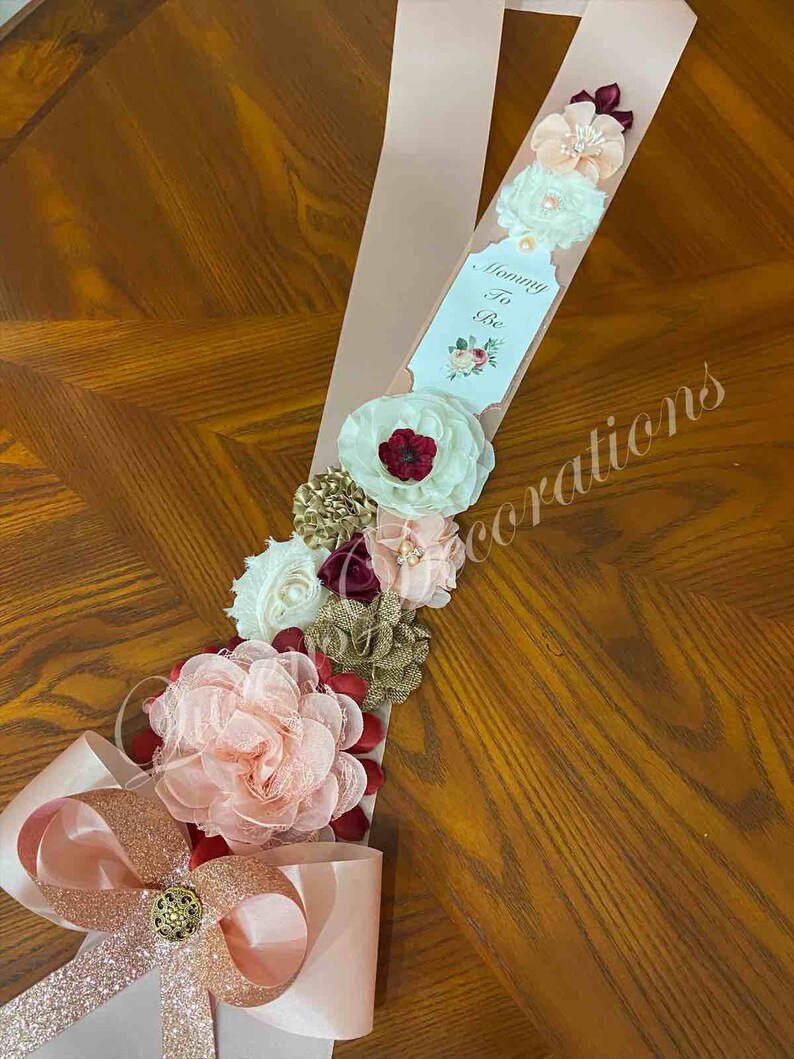 May include: A pink satin sash with a white card that says "Mommy To Be" and a floral design. The sash is decorated with burgundy, blush, and gold flowers.
