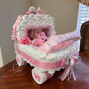 May include: A pink and white nappy cake shaped like a baby carriage with a doll inside. The carriage is decorated with pink and white polka dots, a pink ribbon, and a silver bow.