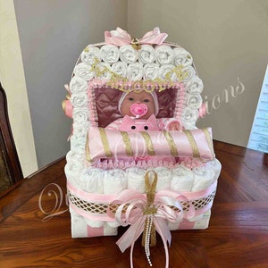 Puede incluir: Un pastel de pa&ntilde;ales rosa y blanco con forma de cochecito de beb&eacute; con una mu&ntilde;eca dentro. El cochecito est&aacute; decorado con una cinta dorada y un lazo rosa.
