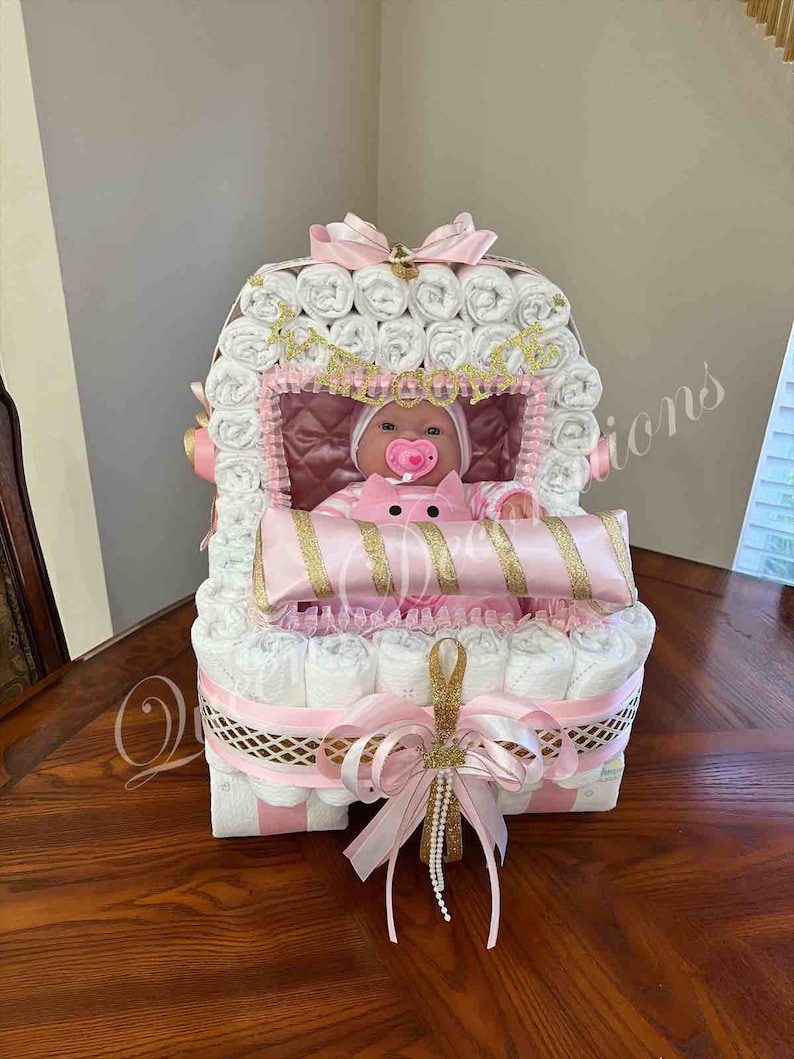 Puede incluir: Un pastel de pa&ntilde;ales rosa y blanco con forma de cochecito de beb&eacute; con una mu&ntilde;eca dentro. El cochecito est&aacute; decorado con una cinta dorada y un lazo rosa.