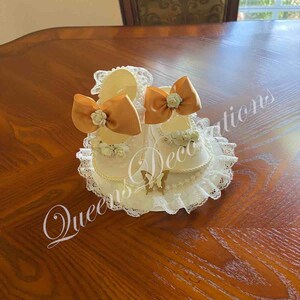 May include: Two white baby shoes with gold bows and lace trim. The shoes are on a white lace base with a gold butterfly.